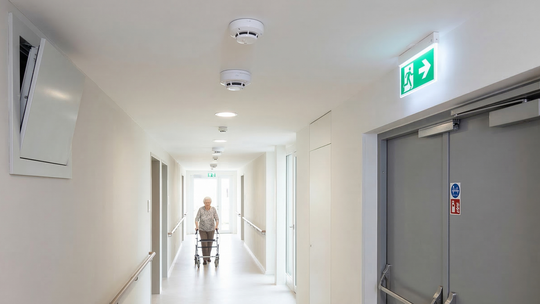 Jasny korytarz w domu opieki z poręczami po obu stronach. Starsza kobieta o balkonik idzie w stronę widza. Na suficie widać czujki dymu. Na lewej ścianie zamontowano dużą klapę systemu oddymiania, a po prawej znajdują się szare drzwi przeciwpożarowe ze świecącym znakiem ewakuacyjnym.