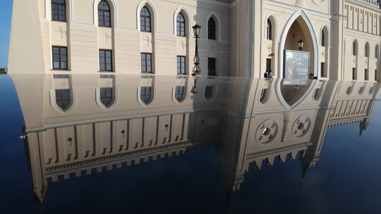 Odbicie fasady neogotyckiego budynku Zamku Lubelskiego w czarnej, lśniącej karoserii samochodu na tle błękitnego nieba.