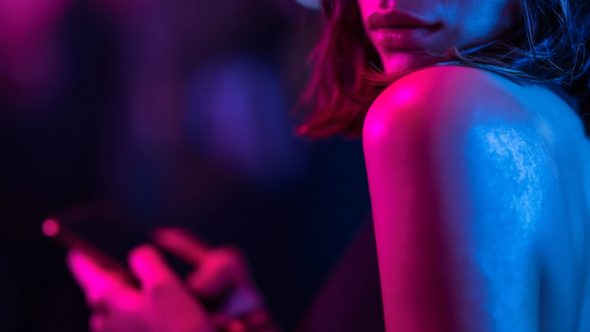 Osoba korzystająca ze smartfona w ciemności, oświetlona różowym i niebieskim światłem neonowym, symbolizująca komunikację cyfrową i poszukiwanie relacji w internecie.