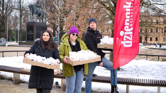Trzy osoby stoją na placu w zimowej scenerii i trzymają kartony z zapakowanymi pączkami. Obok nich stoi czerwony baner „Dziennik Wschodni”