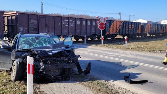 Rozbity samochód osobowy po wypadku na przejeździe kolejowym – uszkodzony pojazd stoi obok torów, gdy przejeżdża pociąg towarowy.