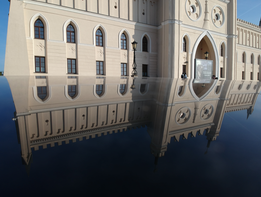 Odbicie fasady neogotyckiego budynku Zamku Lubelskiego w czarnej, lśniącej karoserii samochodu na tle błękitnego nieba.