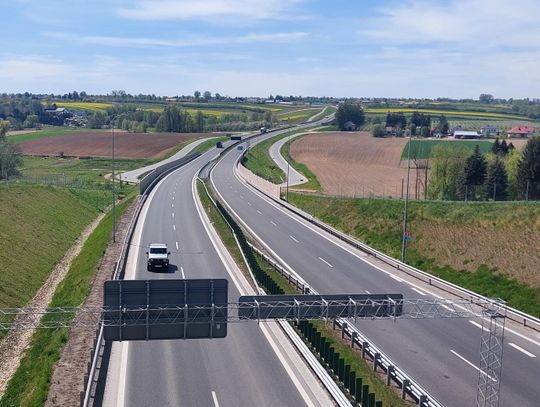 Na zdjęciu widoczna jest droga krajowa przebiegająca przez tereny rolnicze. Jezdnią poruszają się samochody ciężarowe i osobowe, a po obu stronach drogi rozciągają się pola uprawne, w tym kwitnący rzepak. Zdjęcie ilustruje inwestycje drogowe, które otrzymają wsparcie finansowe z Europejskiego Banku