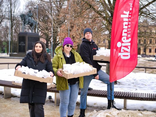 Trzy osoby stoją na placu w zimowej scenerii i trzymają kartony z zapakowanymi pączkami. Obok nich stoi czerwony baner „Dziennik Wschodni”