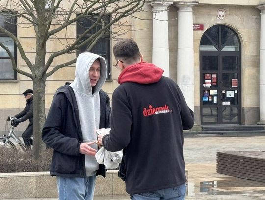 Dziennikarz Dziennika Wschodniego z torebkami wypełnionymi pączkami wręcza drugiemu mężczyźnie paczkę z pączkiem