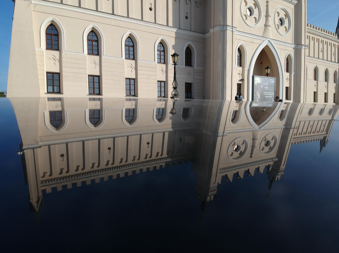 Odbicie fasady neogotyckiego budynku Zamku Lubelskiego w czarnej, lśniącej karoserii samochodu na tle błękitnego nieba.