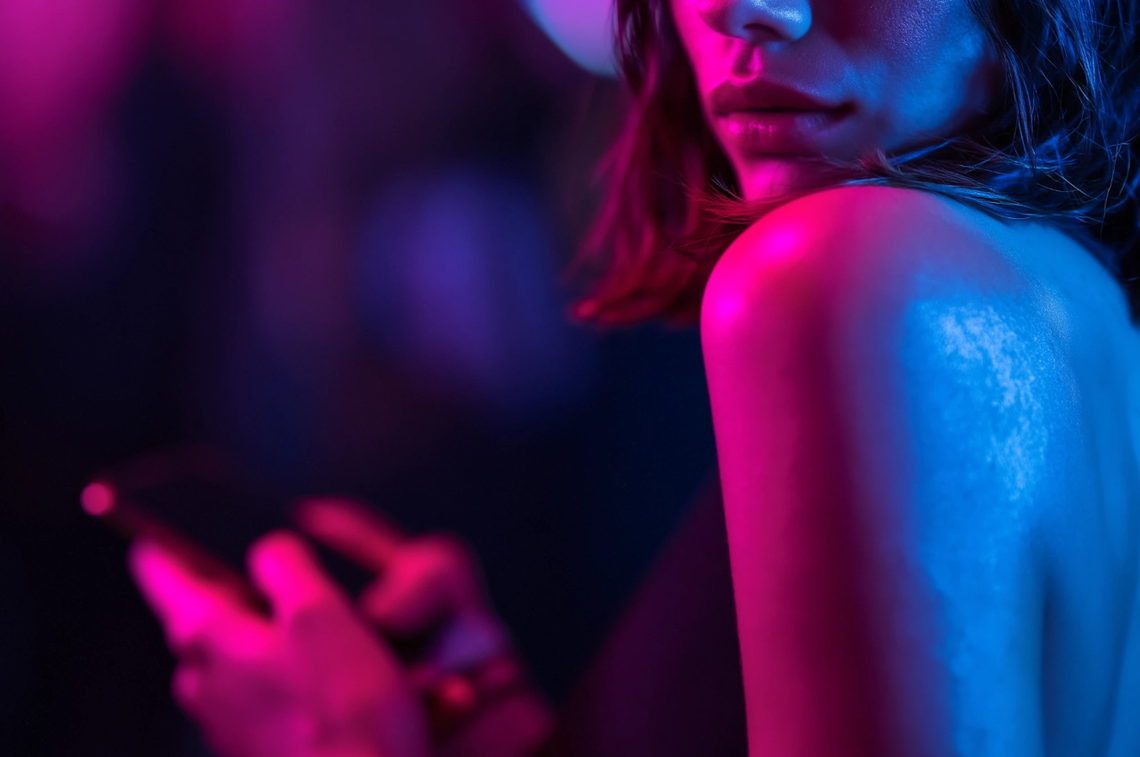 Od samotności do dialogu: jak internet pomaga znaleźć przyjaciół i wsparcie Osoba korzystająca ze smartfona w ciemności, oświetlona różowym i niebieskim światłem neonowym, symbolizująca komunikację cyfrową i poszukiwanie relacji w internecie.