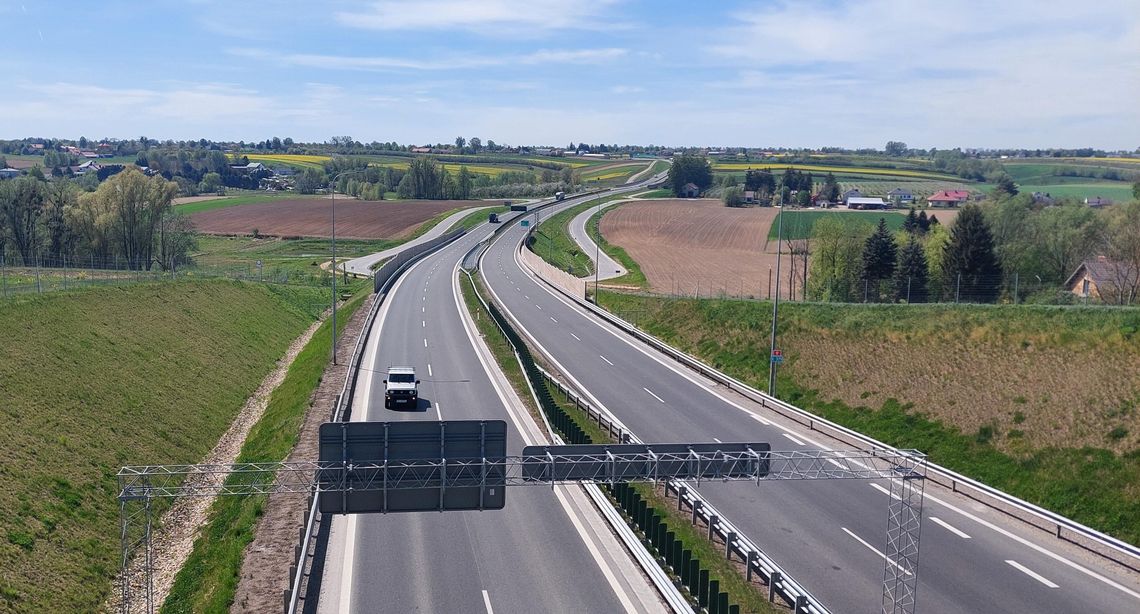 Na zdjęciu widoczna jest droga krajowa przebiegająca przez tereny rolnicze. Jezdnią poruszają się samochody ciężarowe i osobowe, a po obu stronach drogi rozciągają się pola uprawne, w tym kwitnący rzepak. Zdjęcie ilustruje inwestycje drogowe, które otrzymają wsparcie finansowe z Europejskiego Banku