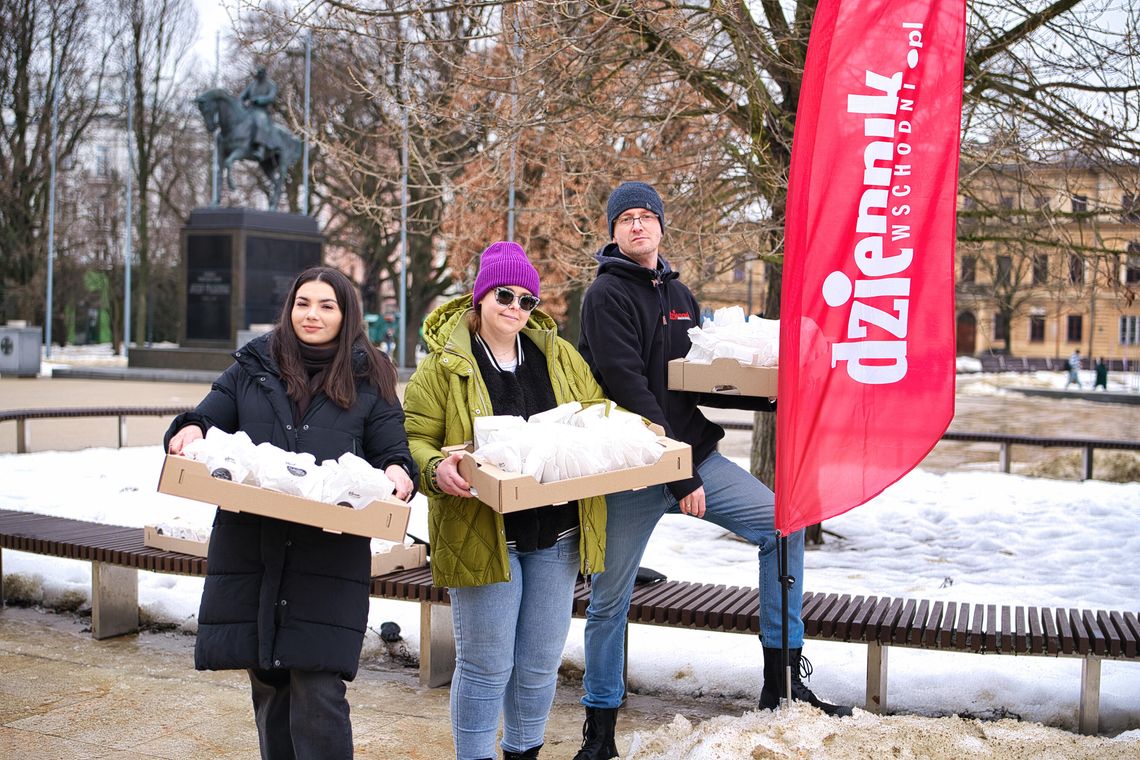 Trzy osoby stoją na placu w zimowej scenerii i trzymają kartony z zapakowanymi pączkami. Obok nich stoi czerwony baner „Dziennik Wschodni”