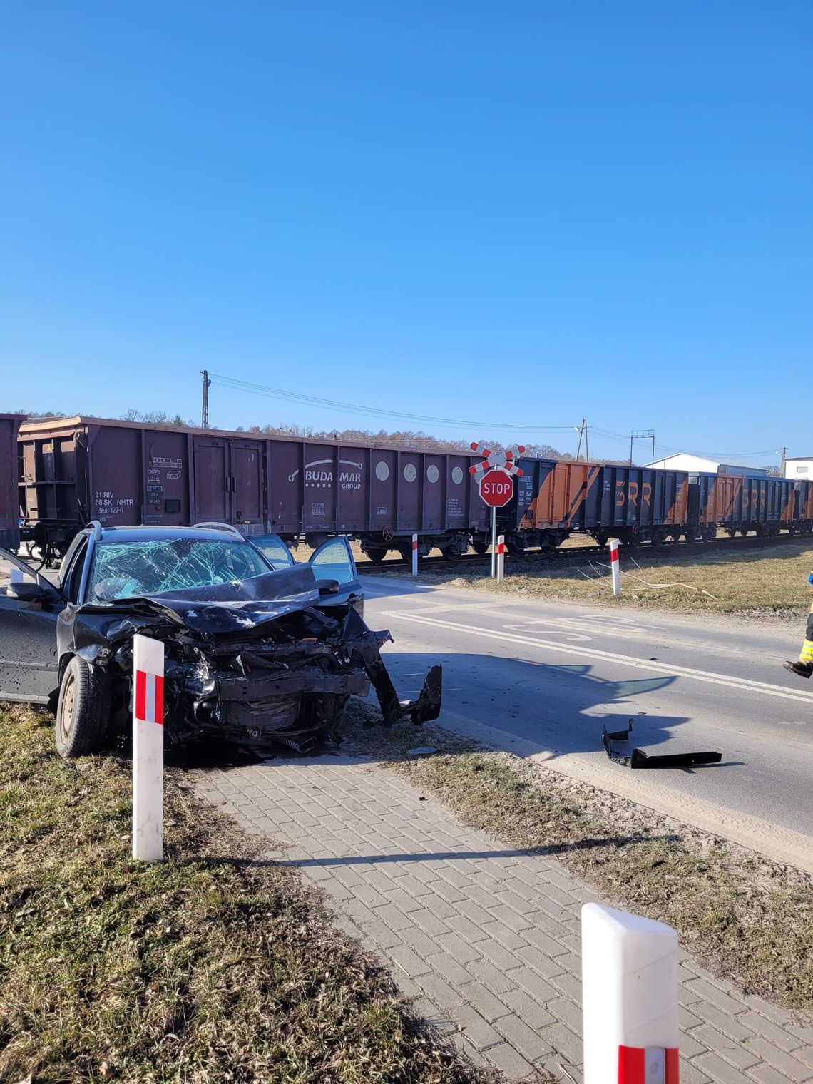 Rozbity samochód osobowy po wypadku na przejeździe kolejowym – uszkodzony pojazd stoi obok torów, gdy przejeżdża pociąg towarowy.