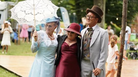 Uczestnicy pikniku w stylu retro w eleganckich strojach z parasolką spacerują po parku podczas letniego wydarzenia.