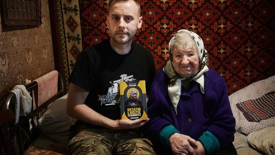 Mężczyzna i starsza kobieta siedzą w domu, prezentując książkę „Ostatni ludzie Czarnobyla” o mieszkańcach strefy wykluczenia.