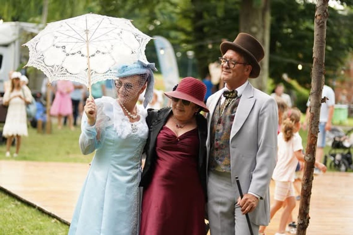 Uczestnicy pikniku w stylu retro w eleganckich strojach z parasolką spacerują po parku podczas letniego wydarzenia.